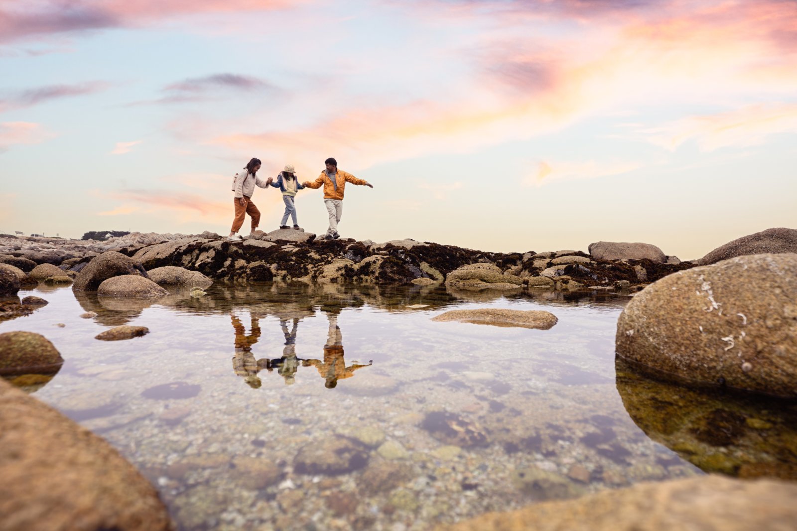 This is an image of a family of three, mom, dad and daughter, holding hands as they carefully cross over rocks to a tide pool at Asilomar State Beach in Pacific Grove, California