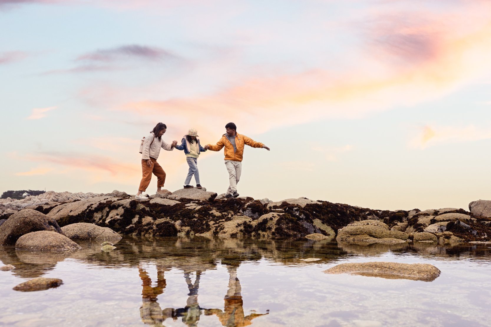 This is an image of a family of three, mom, dad and daughter, holding hands as they carefully cross over rocks to a tide pool at Asilomar State Beach in Pacific Grove, California