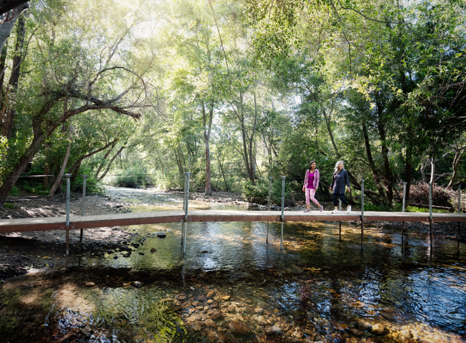 This is an image of two women hiking across a bridge over a river at Garland Ranch Regional Park in Carmel Valley. The women are surrounded by greenery and trees