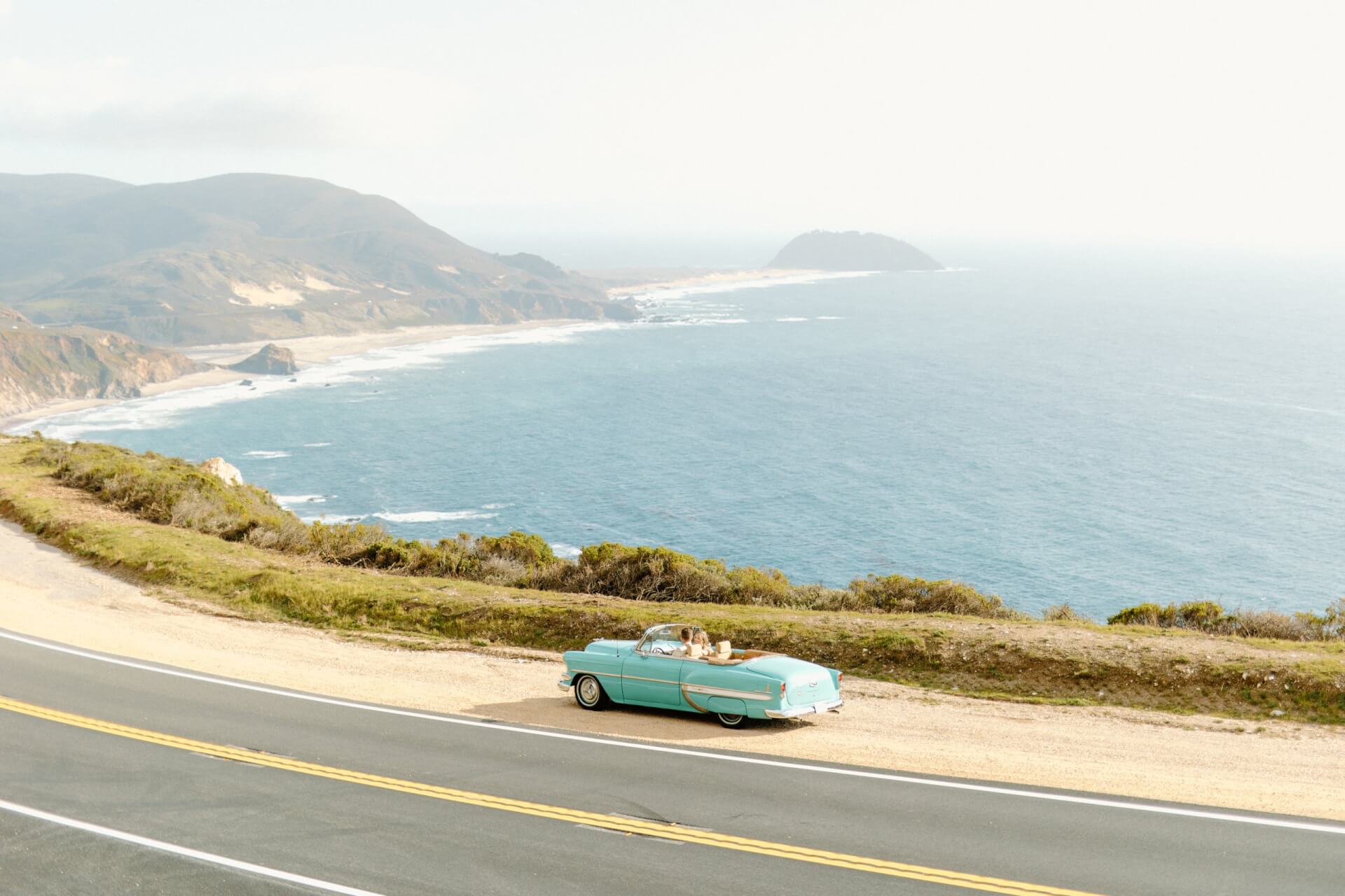 This is an image of a blue classic car pulled over in a car turn out on Highway 1 with the Big Sur coastline in the background