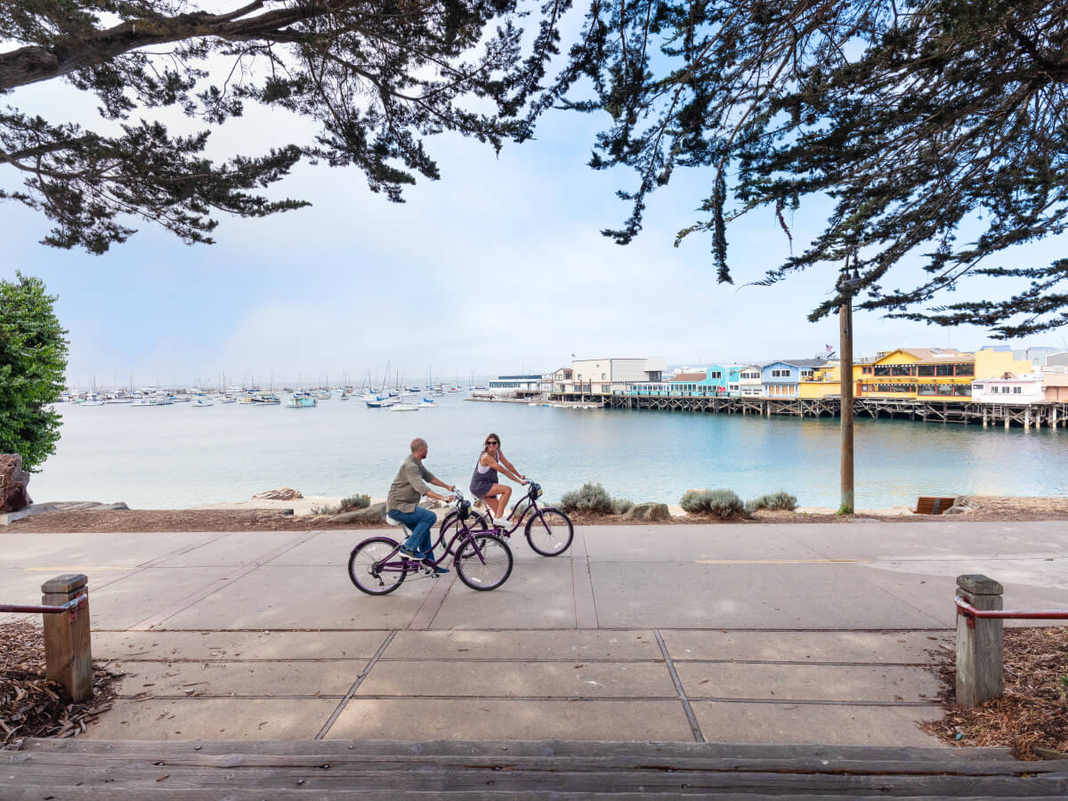 This is an image of a man and woman riding bicycles on a sidewalk on the Monterey Bay Coastal Recreation Trail. The colorful buildings of Old Fisherman's Wharf can be seen int he background, as well as the ocean lined with sailboats