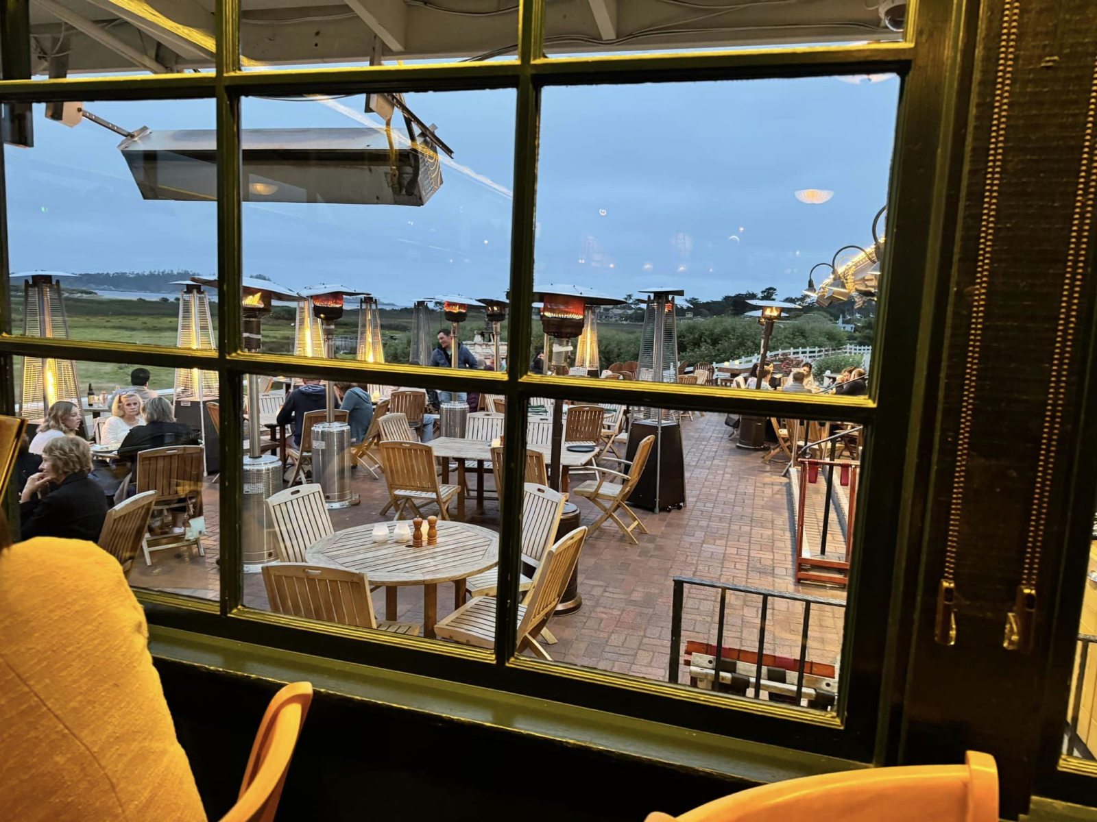 This is an image of the outdoor patio tables at the restaurant at Carmel Mission Ranch in Carmel Valley taken from an indoor table. The wooden tables and chairs are surrounded by heat lamps, creating a warm ambiance. The ocean can be seen from a distance.