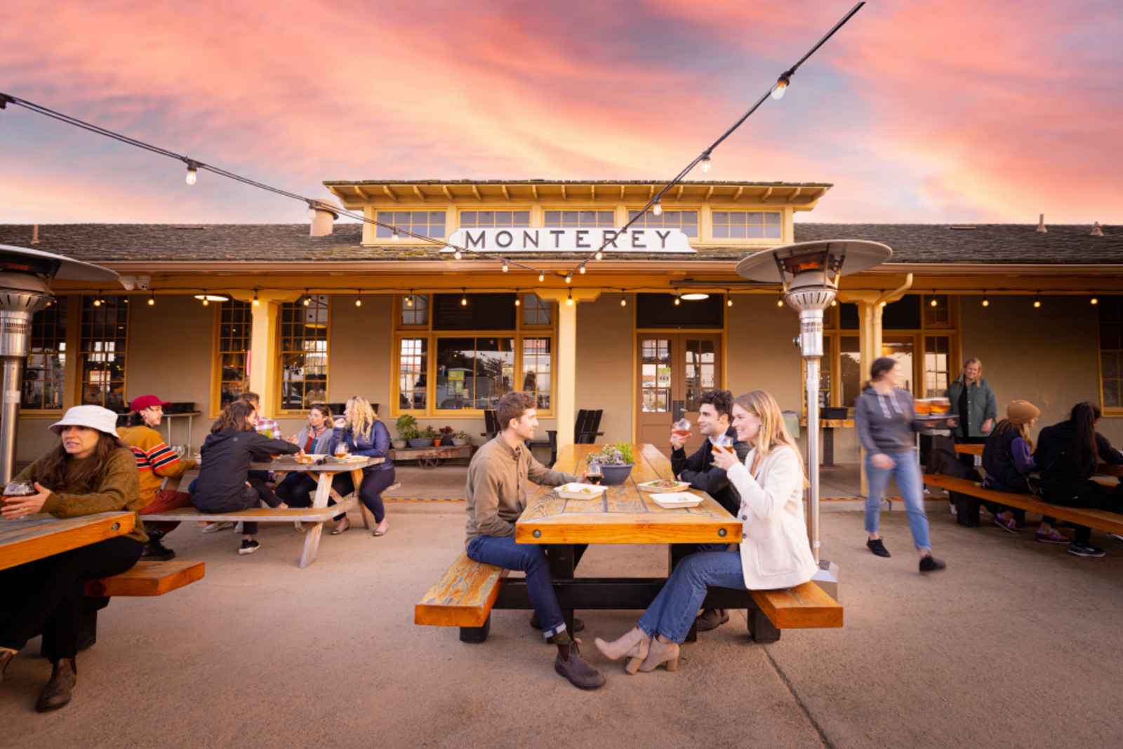 This is an image of the exterior of Dust Bowl Brewing Co. in Monterey at sunset. Various groups of friends sit at wooden bench style seating in the breweries outdoor area, laughing and drinking beer together.