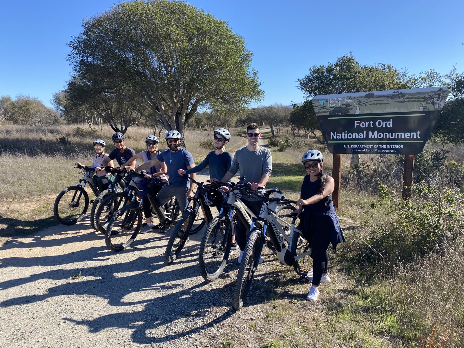 This is an image of a group of friends posing in front of the Fort Ord National Momnument sign with their eBikes and helmets during a VineCycle eBike Tour