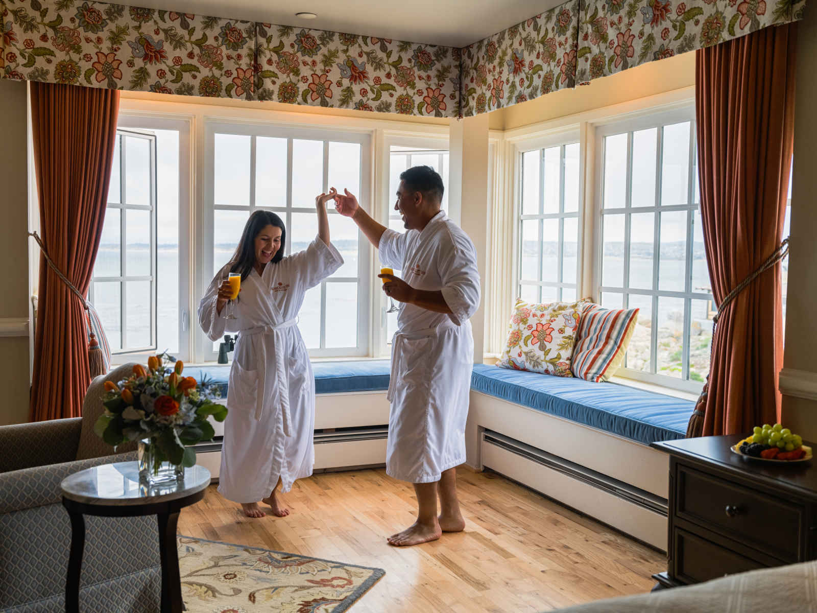 This is an image of a man and a woman in their ocean-view hotel room at Spindrift Inn in Monterey, California. The couple are dancing in their robes in front of the ocean-view window, smiling and holding mimosas.