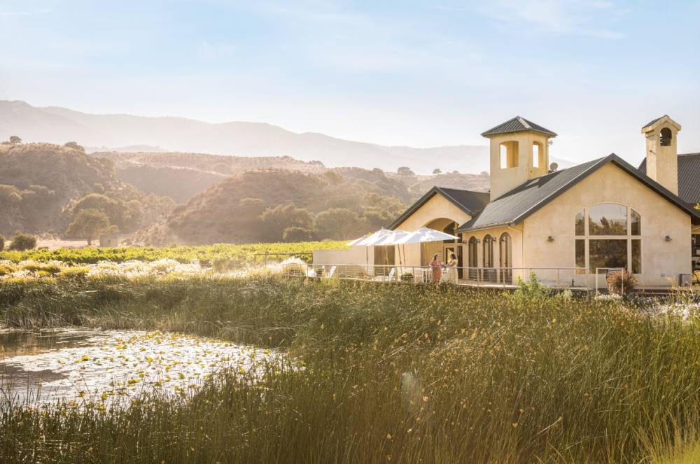 This is an image of Wrath Winery exterior surrounded by a pond, greenery and mountains along River Road in Salinas Valley